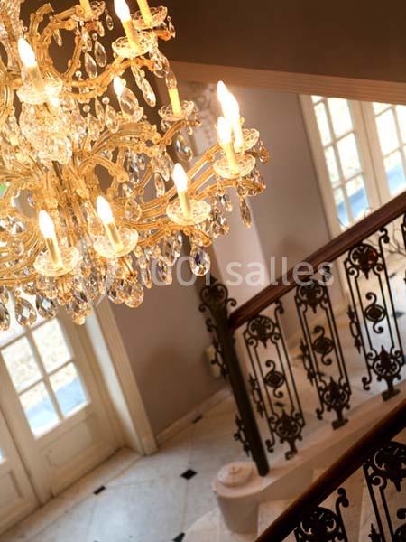 Lustre en cristal éclairé suspendu au-dessus d'un escalier en colimaçon avec rampe en fer forgé, près de fenêtres.