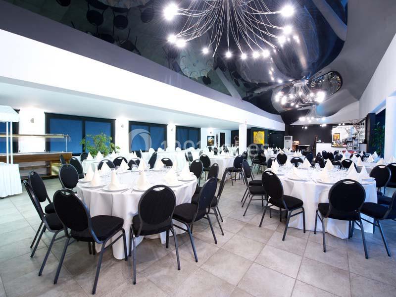 Salle de réception moderne avec tables rondes dressées de nappes blanches, chaises noires et éclairage design.