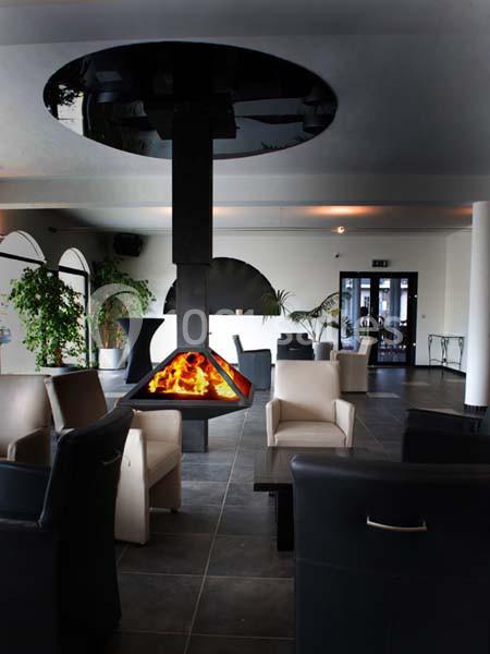 Cheminée suspendue moderne allumée dans un salon spacieux avec fauteuils, plantes et grandes ouvertures lumineuses.