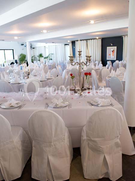 Salle de réception décorée avec des tables rondes et rectangulaires, nappes blanches et chandeliers centraux.