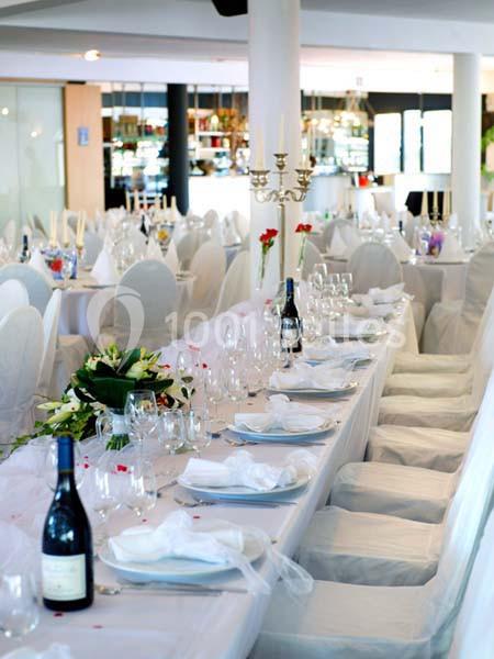 Salle de réception avec tables dressées, nappes blanches, chandeliers, fleurs et bouteilles de vin.