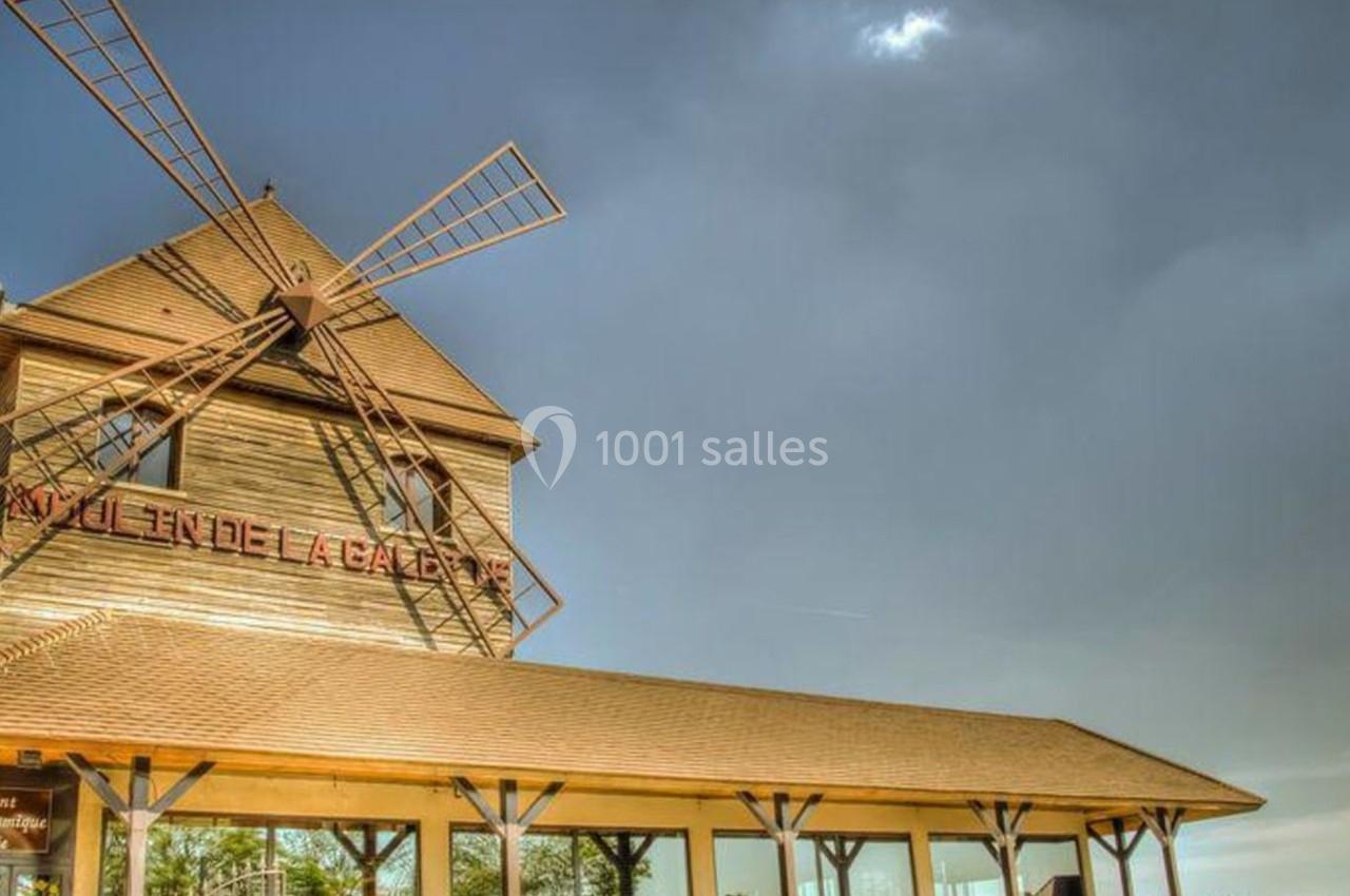 Un moulin en bois avec des ailes fixes, surmontant un bâtiment à toit incliné sous un ciel nuageux.