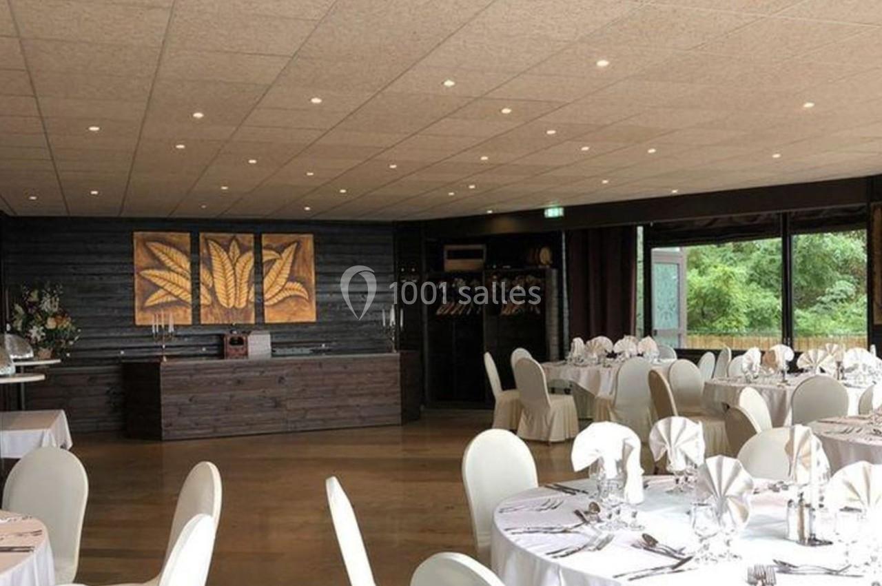 Salle de réception lumineuse avec tables rondes dressées, nappes blanches et grandes baies vitrées donnant sur un espace…