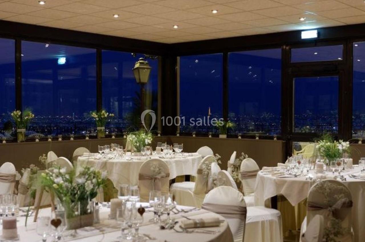 Salle de réception élégante avec tables décorées, grandes baies vitrées offrant une vue nocturne sur une ville illuminée.