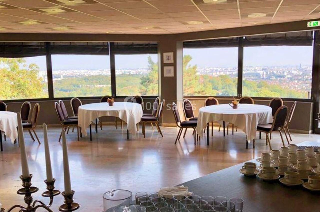 Salle lumineuse avec grandes baies vitrées, tables rondes dressées et vue panoramique sur un paysage urbain et verdoyant.