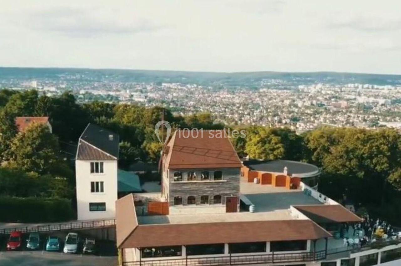 Vue aérienne d'un bâtiment en pierre et bois entouré de verdure, avec une ville visible à l'arrière-plan.