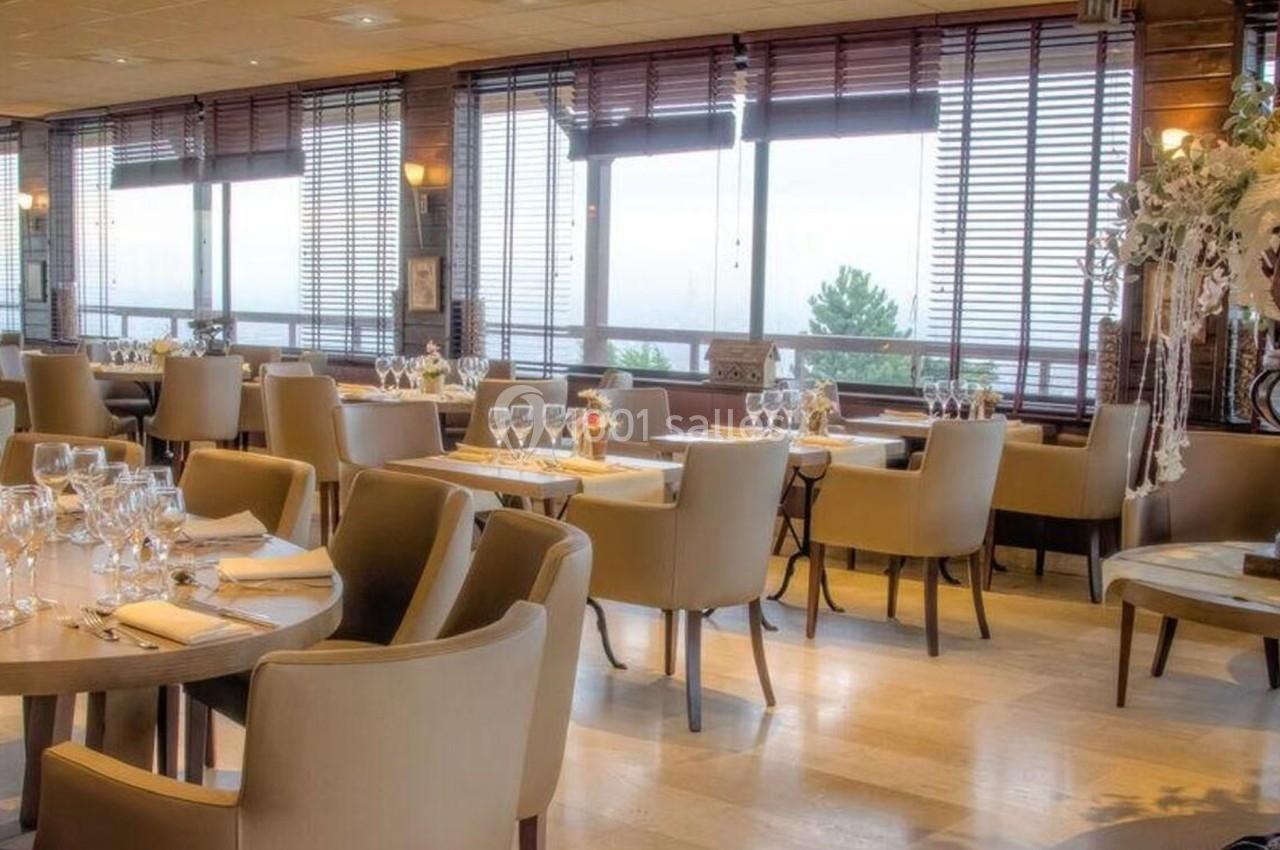 Salle de restaurant élégante avec tables dressées, chaises beige clair et grandes fenêtres laissant entrer la lumière…