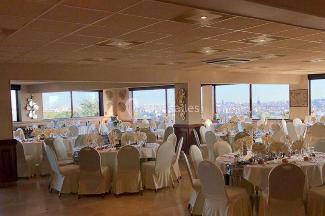 Salle de réception lumineuse avec tables rondes dressées, nappes blanches et vue panoramique sur une ville.