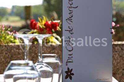 Menu posé sur une table dressée en extérieur, entourée de verres, fleurs et paysage verdoyant en arrière-plan.