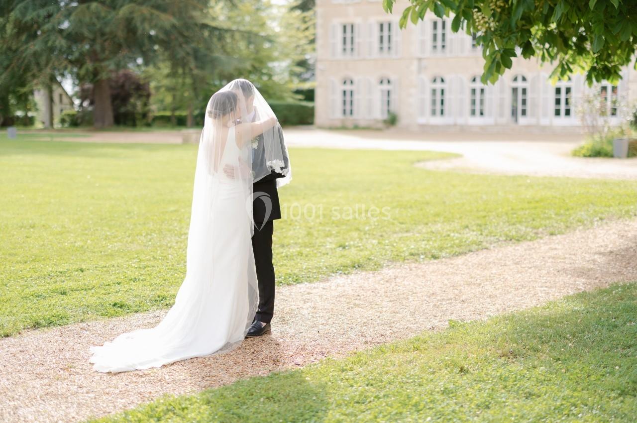 Un couple de mariés s'embrasse sous un voile dans un jardin, près d'un bâtiment en pierre avec de grandes fenêtres.