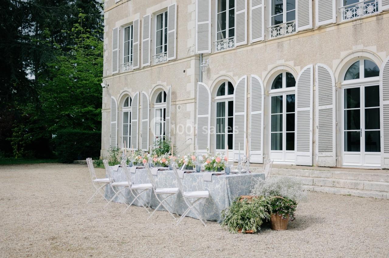 Table dressée en extérieur avec des chaises blanches devant une grande bâtisse aux volets ouverts.