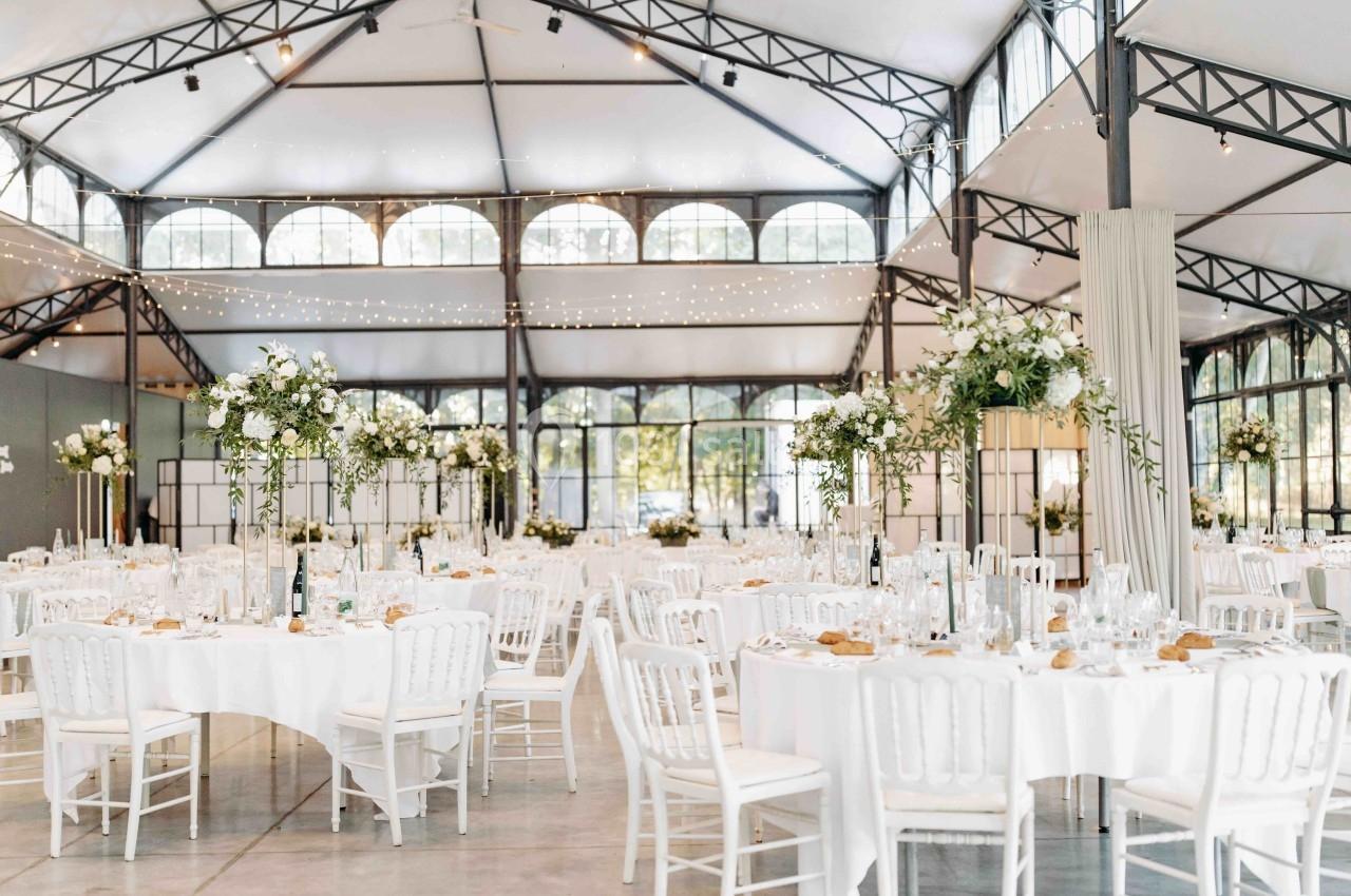 Salle de réception lumineuse avec tables rondes dressées, décorations florales et guirlandes lumineuses suspendues.