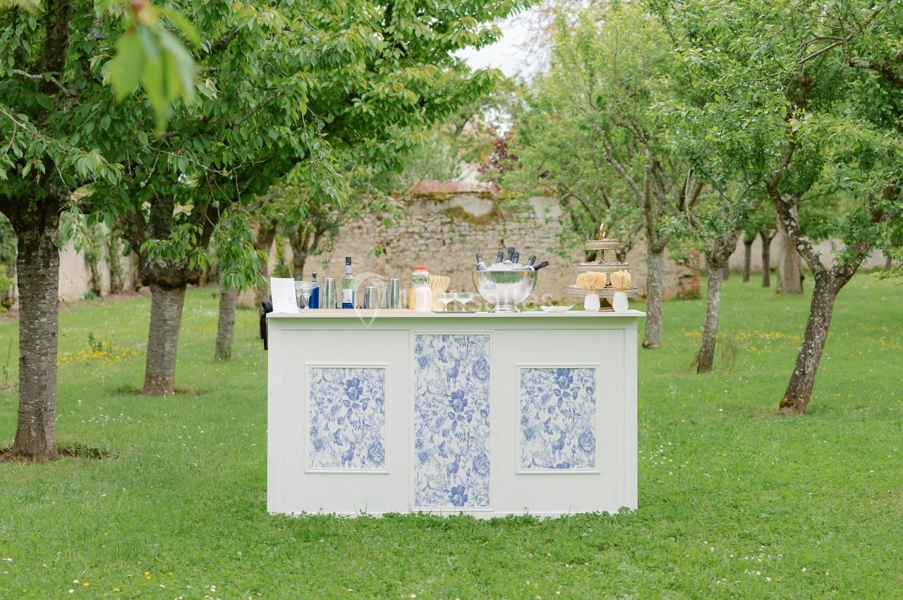 Bar extérieur blanc décoré de motifs bleus, installé dans un jardin verdoyant avec des arbres et des boissons disposées…