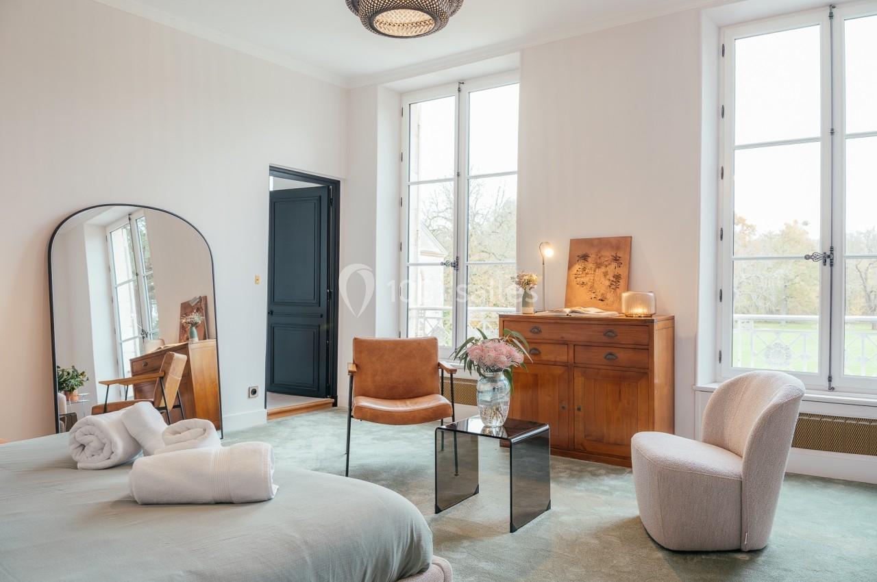 Chambre lumineuse avec lit double, fauteuils, commode en bois, miroir et grandes fenêtres donnant sur un jardin.