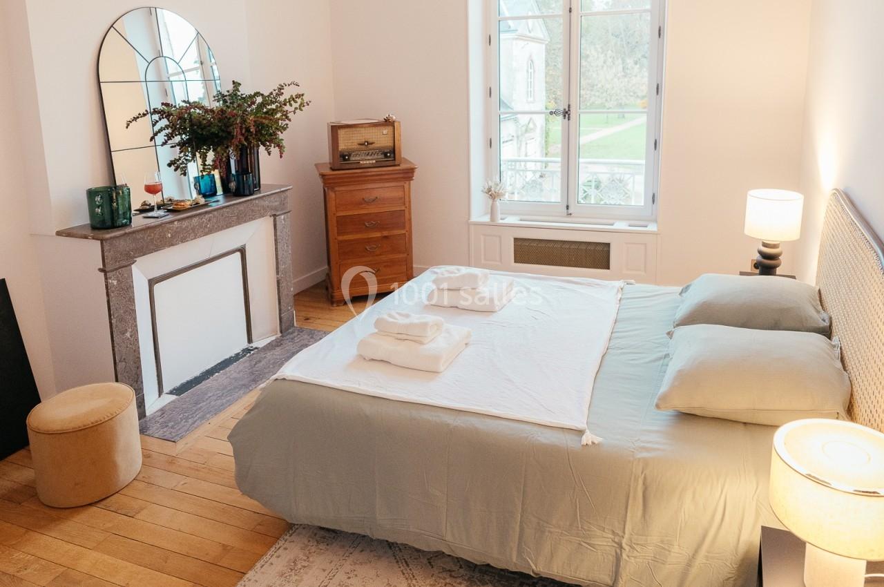 Chambre lumineuse avec lit double, parquet, cheminée décorative, miroir, et vue sur un jardin par une grande fenêtre.