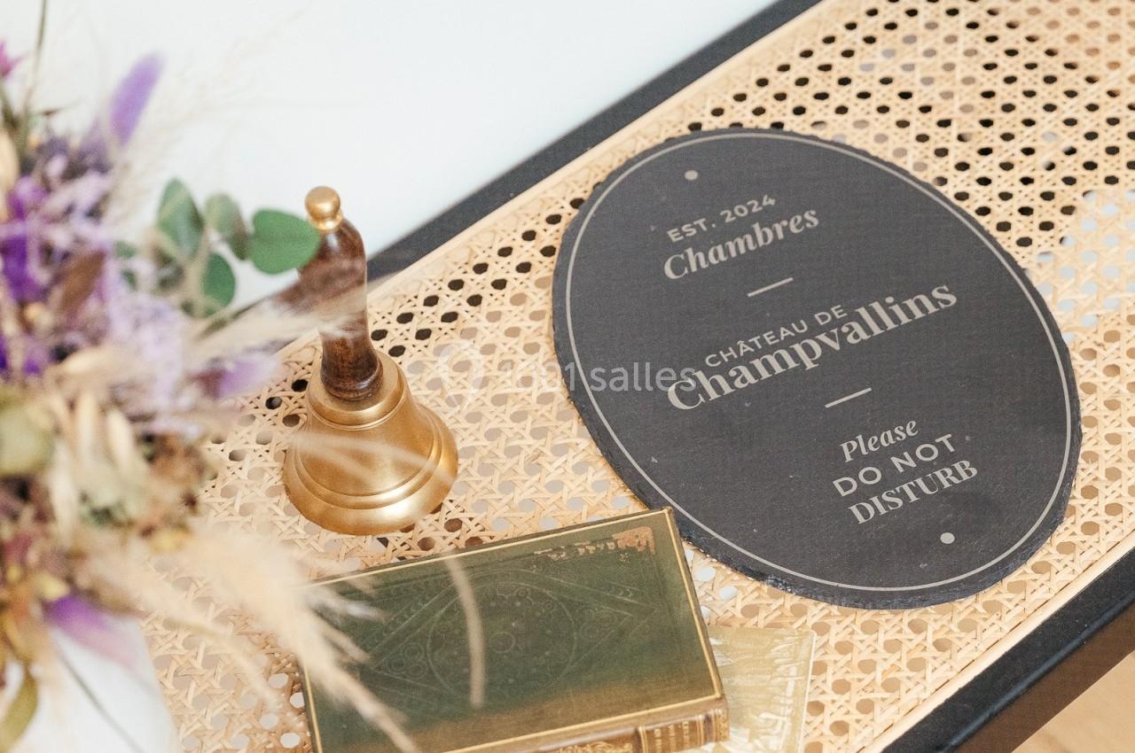 Plaque ’Château de Champvallins’ sur une table en rotin avec une cloche dorée et une boîte décorative.