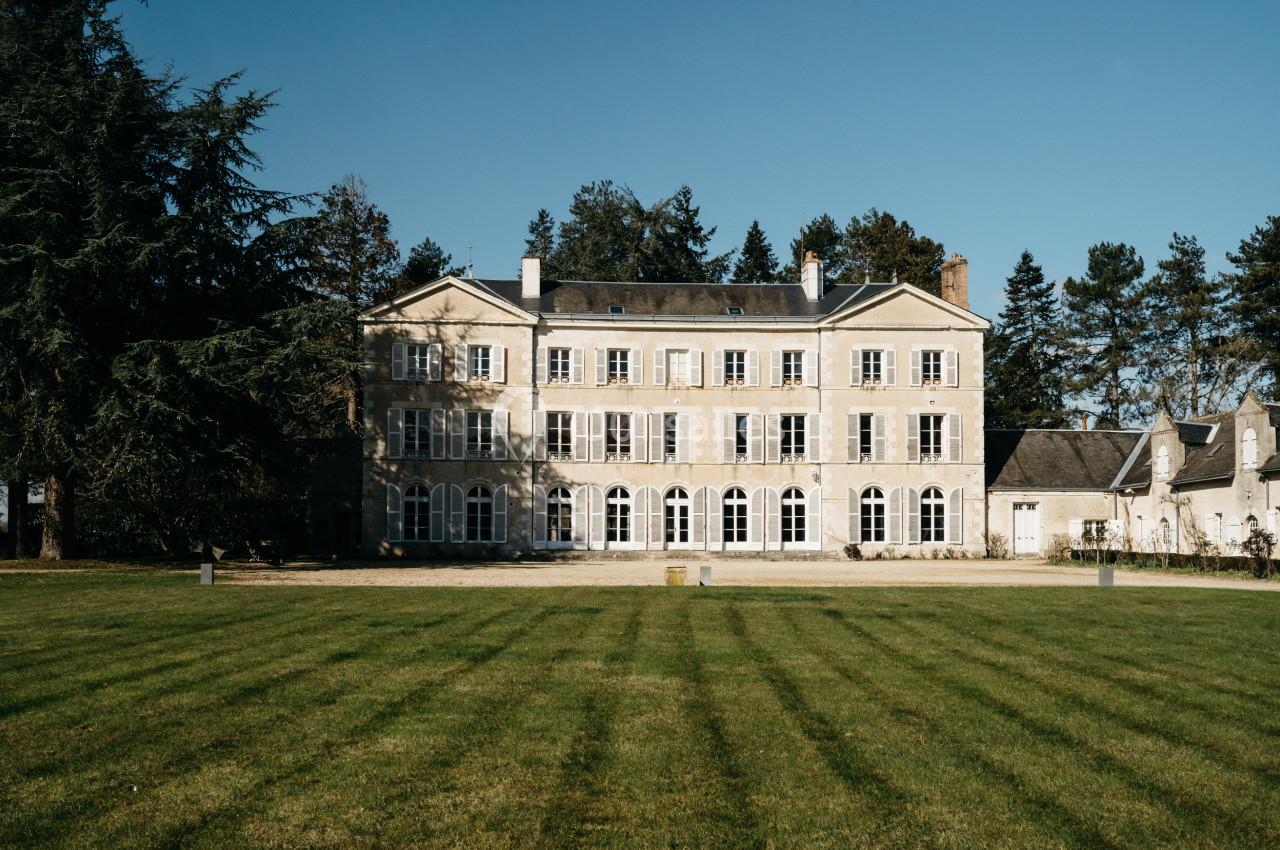 Façade d'un grand bâtiment en pierre avec des volets blancs, entouré d'arbres et donnant sur une pelouse.