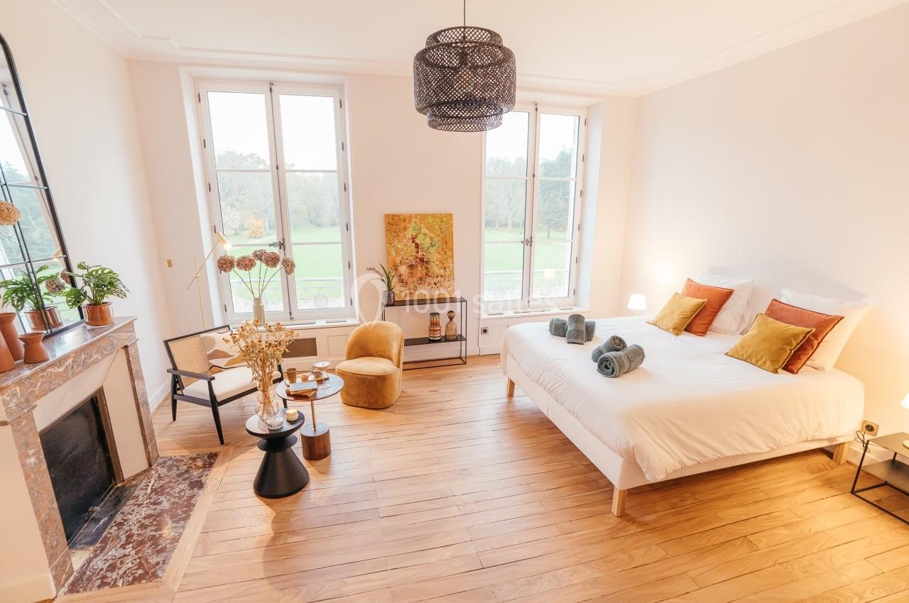 Chambre lumineuse avec lit double, parquet, cheminée, fauteuils, plantes et vue sur un parc à travers de grandes fenêtres.