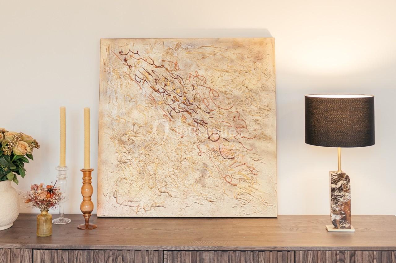 Peinture abstraite beige et dorée accrochée au-dessus d'un buffet en bois, décoré de bougies, fleurs et lampe moderne.