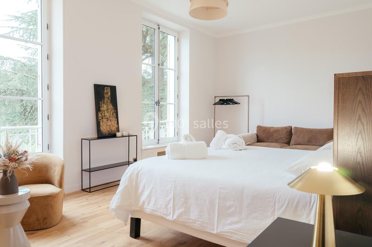 Chambre lumineuse avec lit double, fauteuil, lampes modernes et grande fenêtre donnant sur un espace arboré.