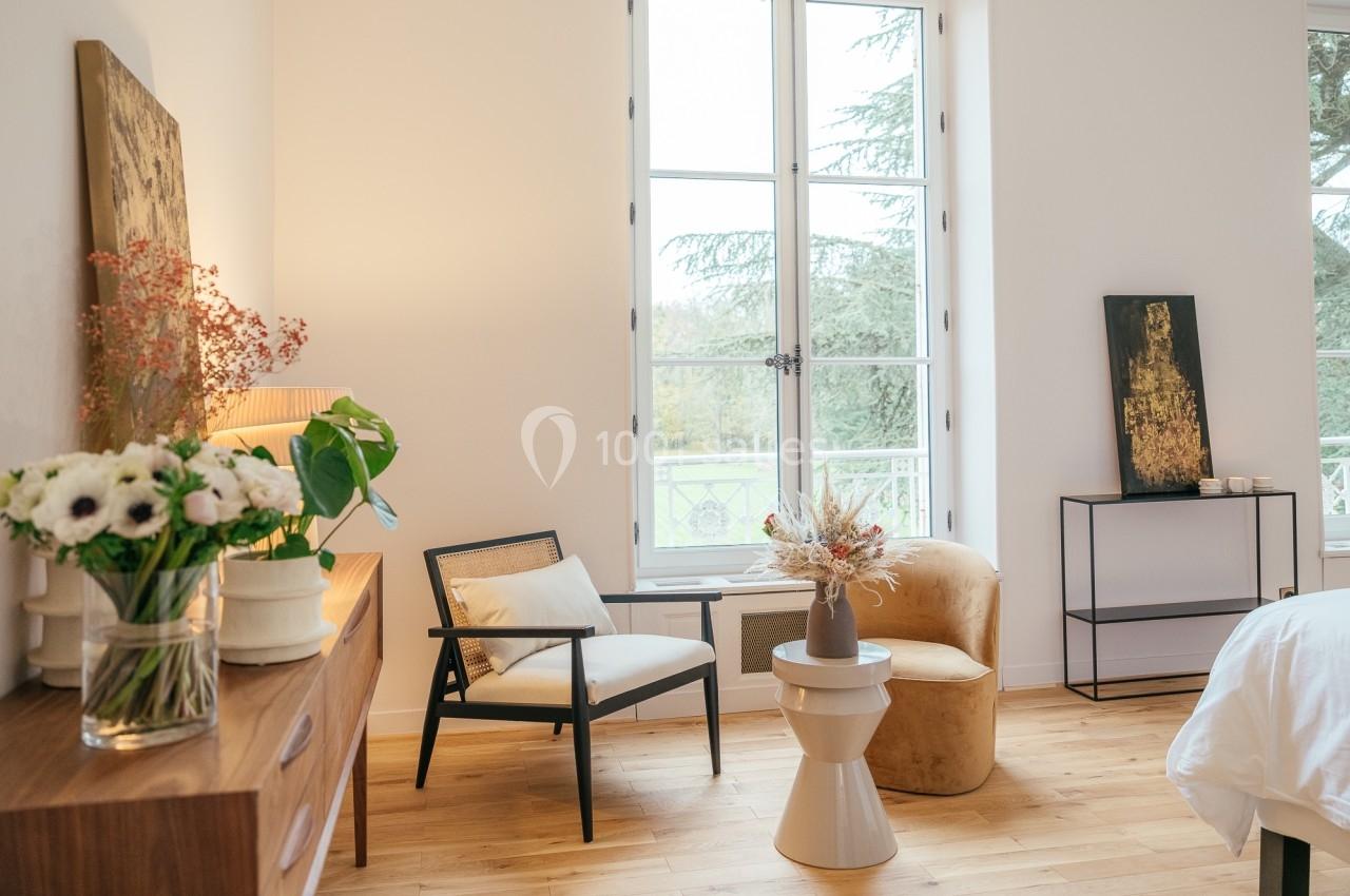 Chambre lumineuse avec fauteuils, table d'appoint, fleurs en vase et grande fenêtre donnant sur un jardin.