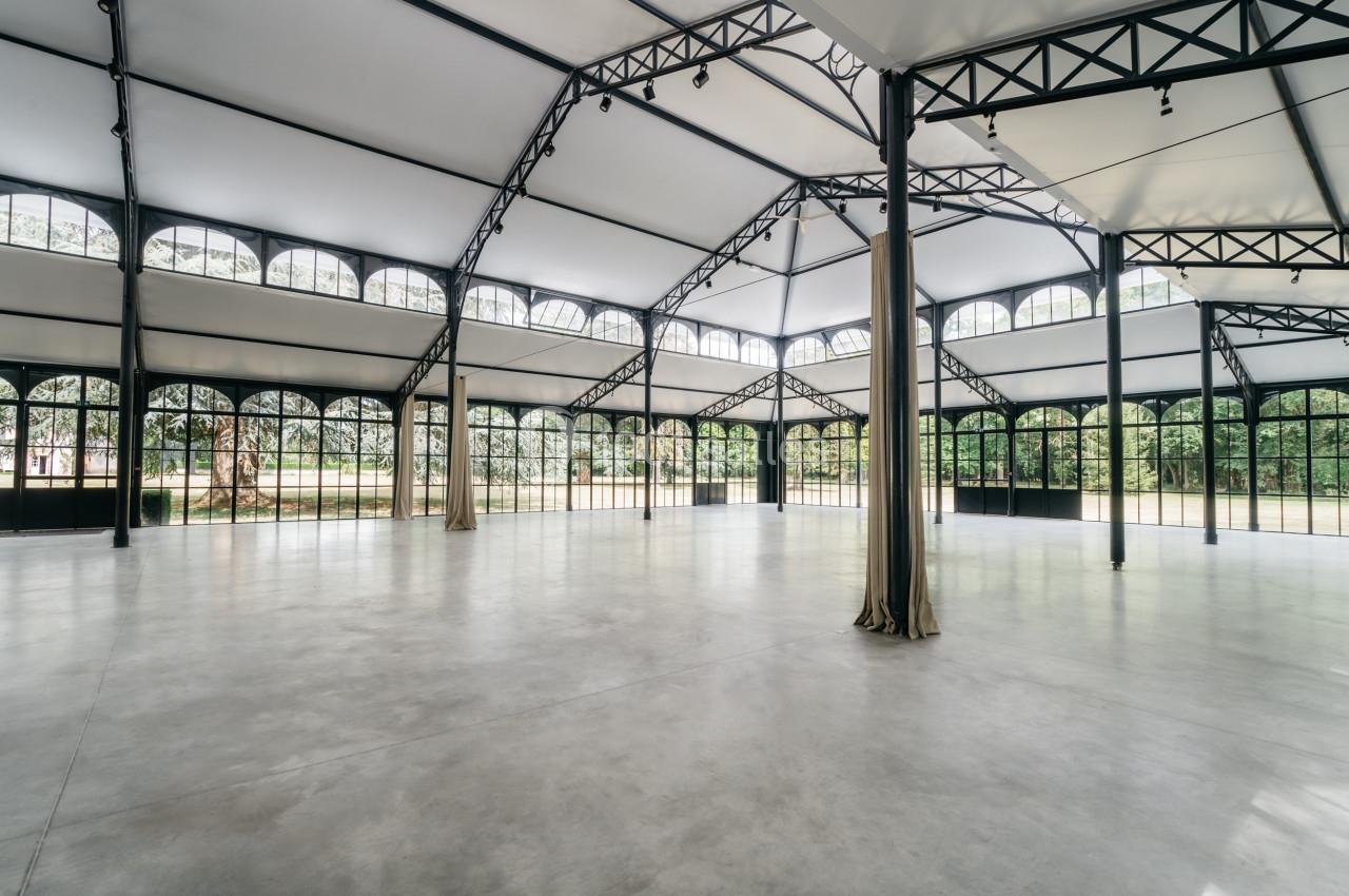 Grande salle lumineuse avec structure métallique, baies vitrées arrondies et sol en béton poli, donnant sur un espace vert.