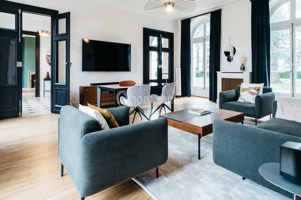 Salon moderne avec canapés bleus, table basse en bois, télévision murale et grandes fenêtres laissant entrer la lumière.