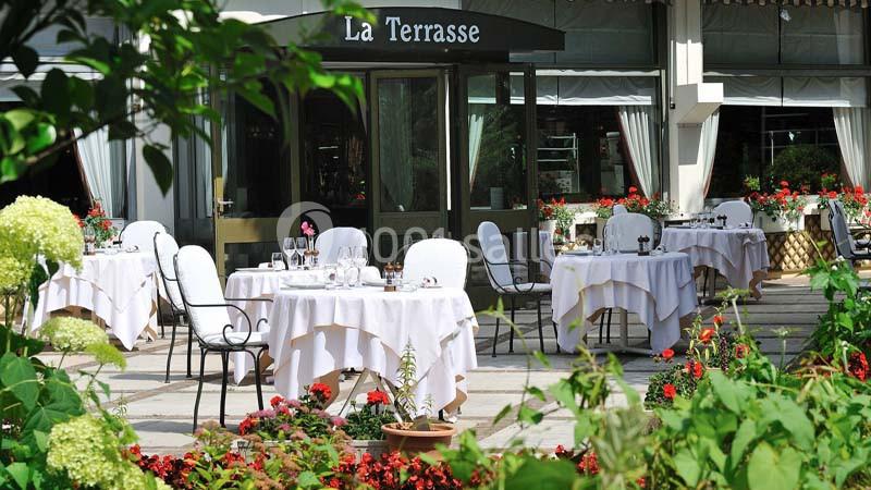 Terrasse d'un restaurant avec tables dressées, nappes blanches et chaises, entourée de fleurs et de verdure.