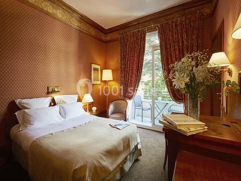 Chambre d'hôtel avec lit double, bureau, fauteuil, balcon donnant sur un espace vert et éclairage chaleureux.