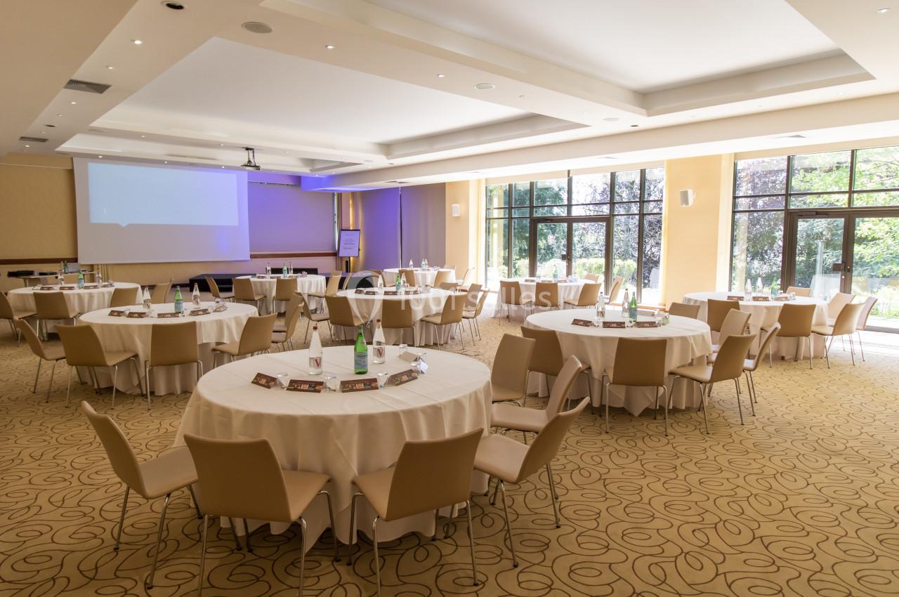 Salle de conférence lumineuse avec des tables rondes dressées, des chaises beige et un écran de projection.