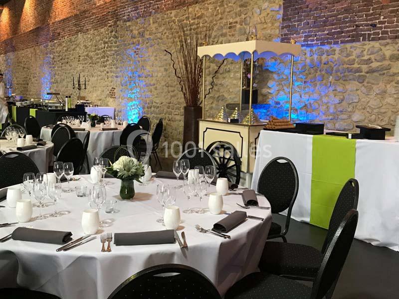 Salle de réception avec tables rondes dressées, nappes blanches, décorations florales et éclairage mural coloré.