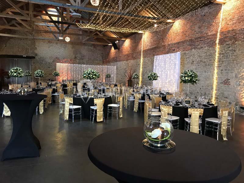 Salle de réception décorée avec des tables rondes, nappes noires, chaises dorées et guirlandes lumineuses suspendues.
