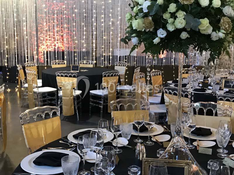 Salle de réception décorée avec des tables dressées, nappes noires, chaises dorées et un grand bouquet floral.