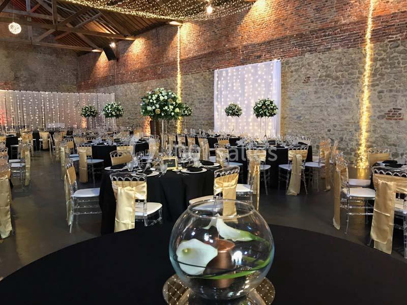 Salle de réception décorée avec des tables rondes, nappes noires, chaises dorées et guirlandes lumineuses suspendues.