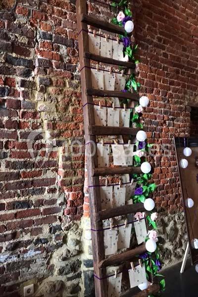 Échelle en bois décorée de fleurs et guirlandes lumineuses, utilisée pour afficher des cartes sur un mur de briques.