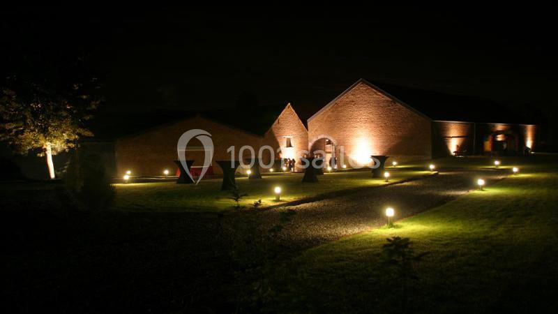 Bâtiment en briques éclairé la nuit, entouré d'un jardin avec allées illuminées par des lampes au sol.