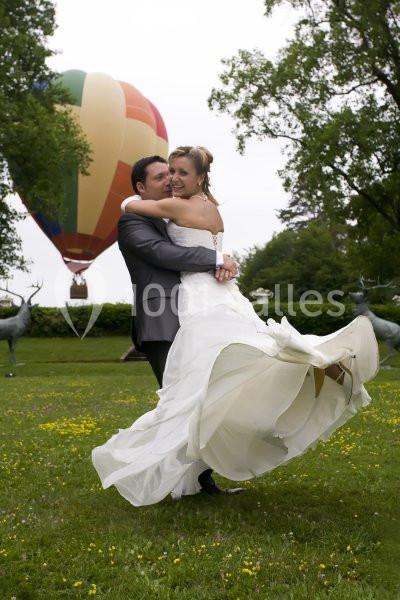Un couple en tenue de mariage s'enlace dans un parc, avec une montgolfière colorée en arrière-plan.