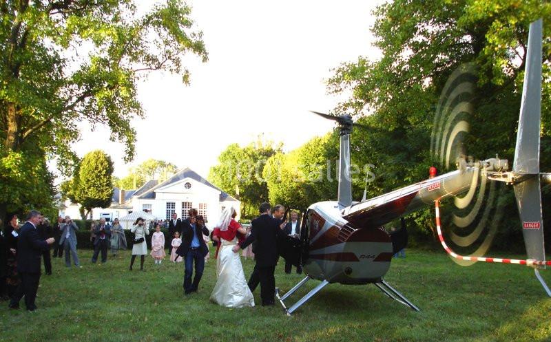 Un couple en tenue de mariage monte à bord d’un hélicoptère dans un jardin entouré d’invités.