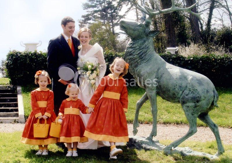 Un couple souriant en tenue de mariage pose avec quatre enfants vêtus de robes orange près d'une statue de cerf.