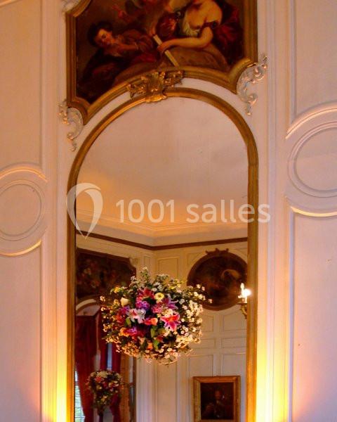 Grand miroir doré reflétant un bouquet de fleurs suspendu, dans une pièce ornée de peintures et de boiseries.