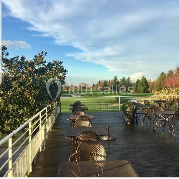 Terrasse en bois avec tables et chaises donnant sur un terrain de golf entouré de végétation sous un ciel dégagé.