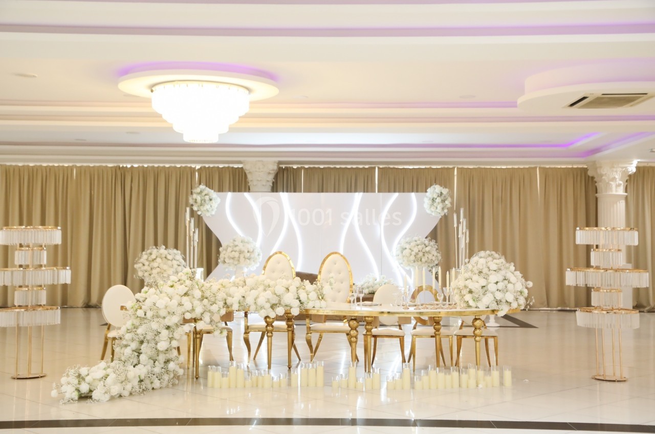 Salle de réception élégante avec décoration florale blanche, chaises dorées et éclairage subtil.