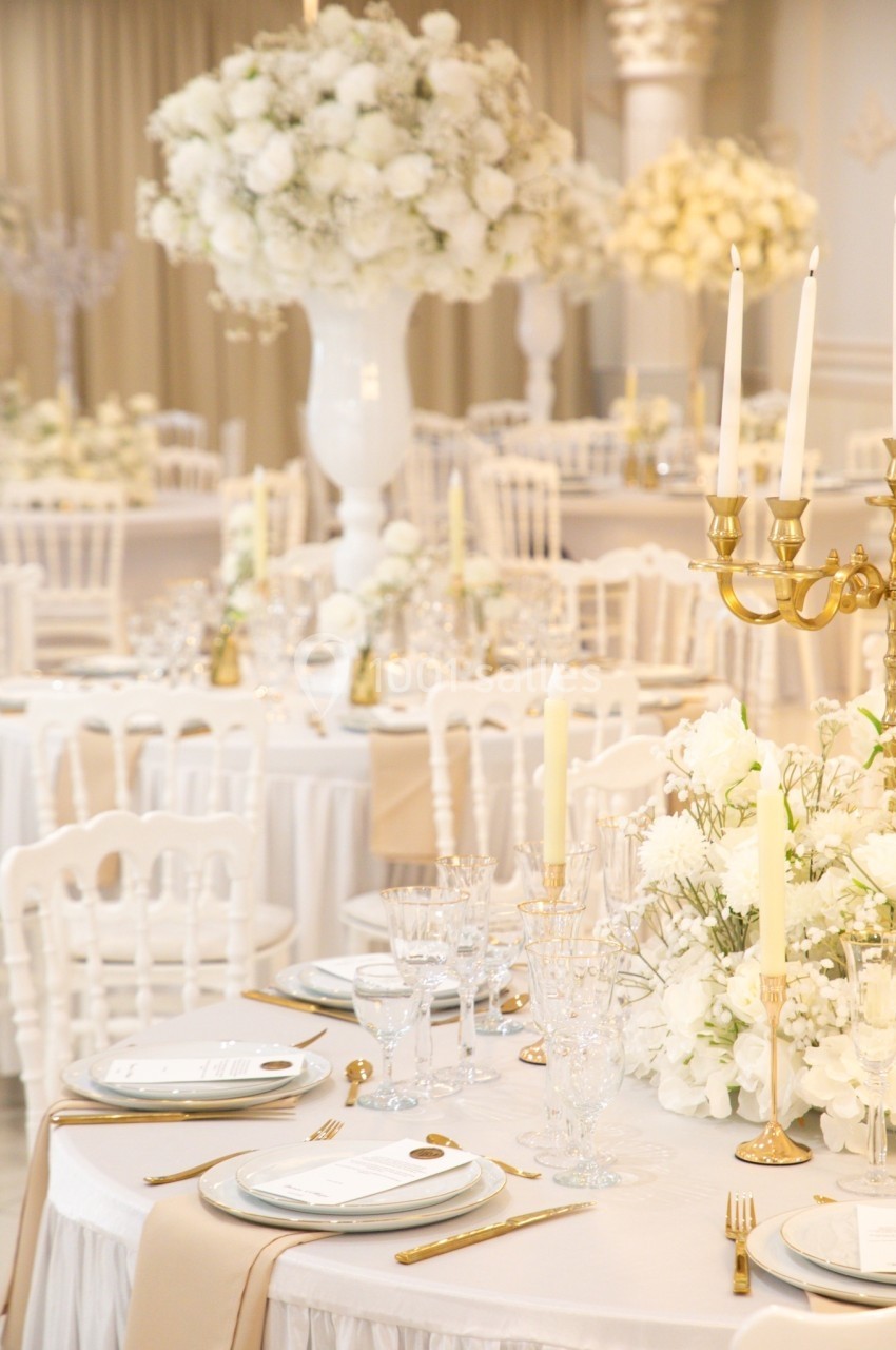 Salle de réception élégante avec tables décorées de fleurs blanches, chandeliers dorés et vaisselle raffinée.