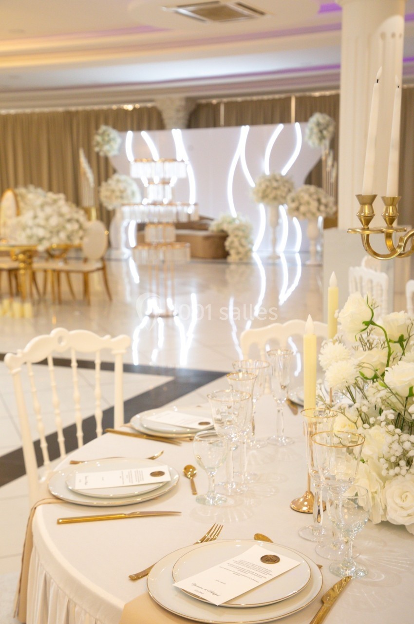 Table élégamment dressée avec vaisselle blanche et dorée, décor floral blanc et arrière-plan de salle de réception lumineuse.