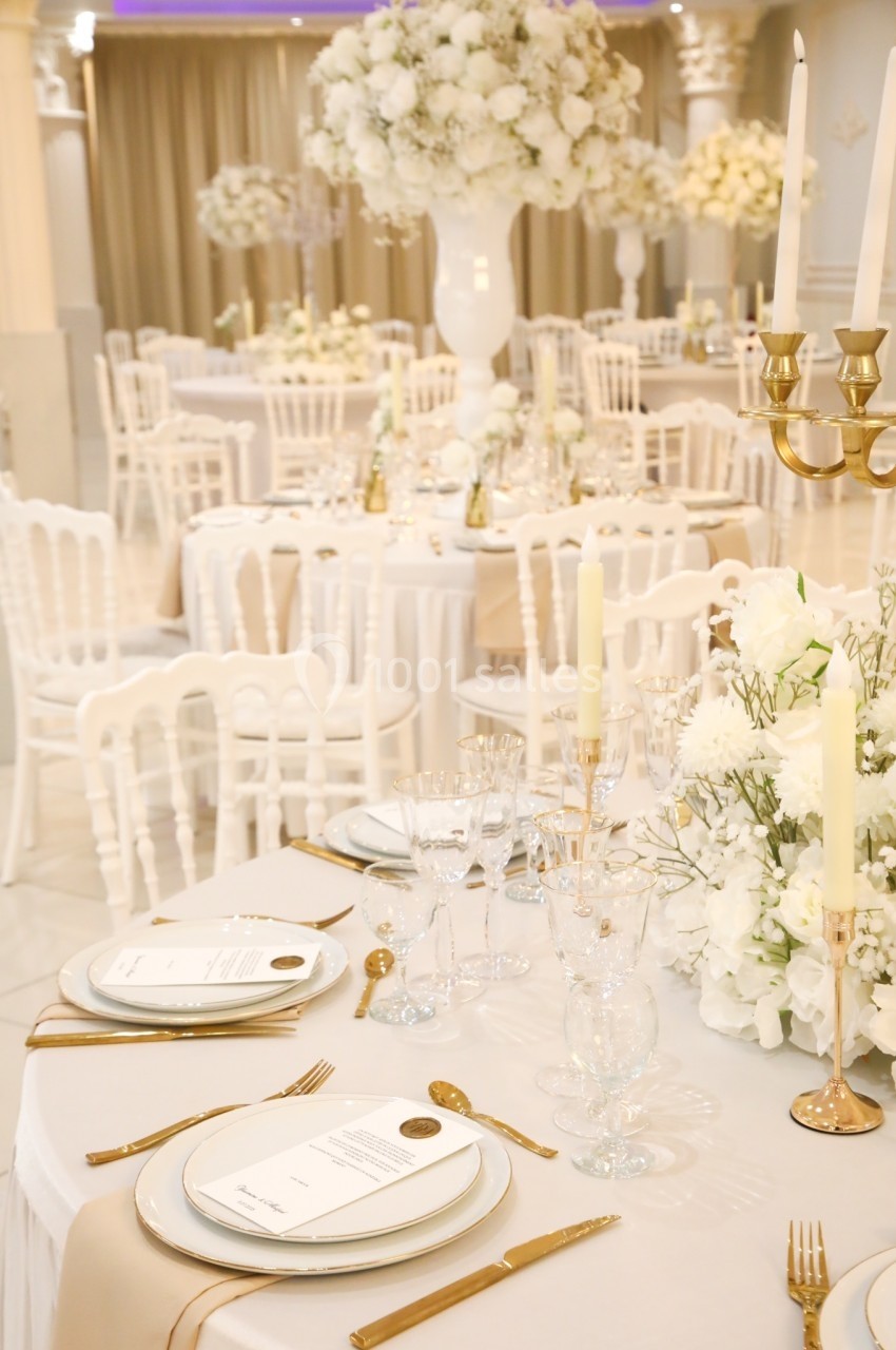 Salle de réception élégante avec tables décorées de nappes blanches, chandeliers dorés et arrangements floraux blancs.