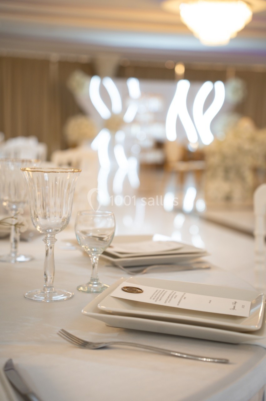Table élégamment dressée avec assiettes blanches, verres en cristal et menu, dans une salle lumineuse au décor raffiné.