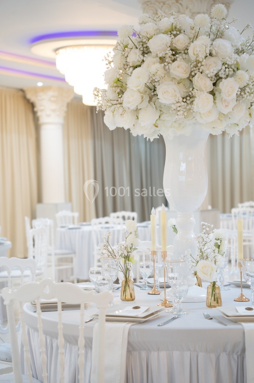 Salle de réception élégante avec tables dressées, nappes blanches et grand centre de table floral blanc.