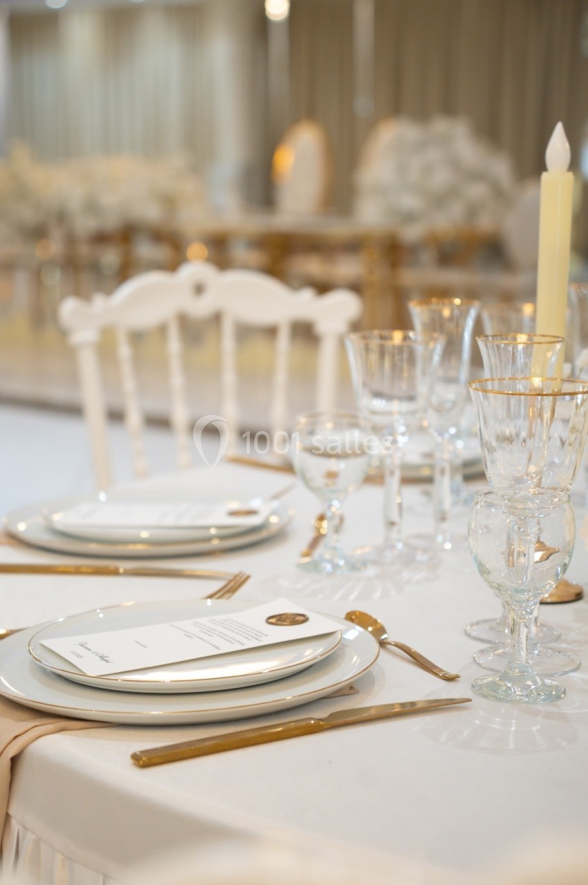 Table élégamment dressée avec assiettes blanches, couverts dorés, verres en cristal et une bougie allumée.