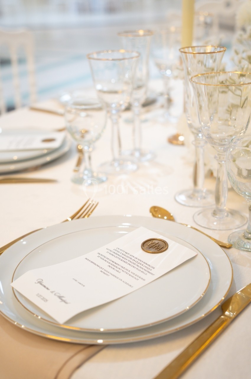 Table élégamment dressée avec assiettes blanches, couverts dorés, verres à pied et menu posé sur une assiette.
