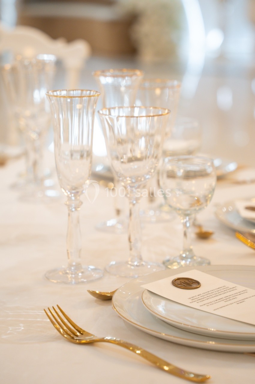 Table élégamment dressée avec vaisselle blanche, couverts dorés et verres à pied, dans un décor lumineux.